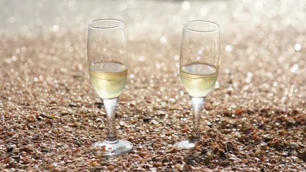 Dwie szklanki szampana stojak na piasku z małe muszle, nad morzem, w fale tło umyć brzegu - Materiał filmowy, wideo