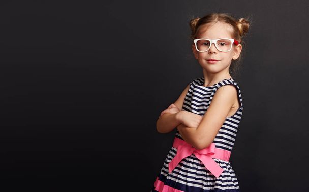 Pretty girl in spectacles standing with crossed arms over black background with copy space. Beautiful smart child at chalkboard. - Фото, зображення