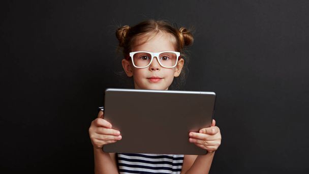 Portrait of smart girl holding digital tablet over black background. Concept of childhood and technology. - Foto, afbeelding