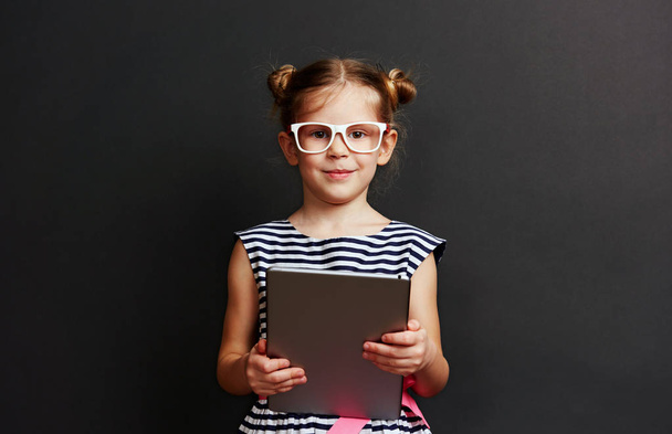 Adorable little girl in glasses holding digital computer on black studio background. - Foto, afbeelding