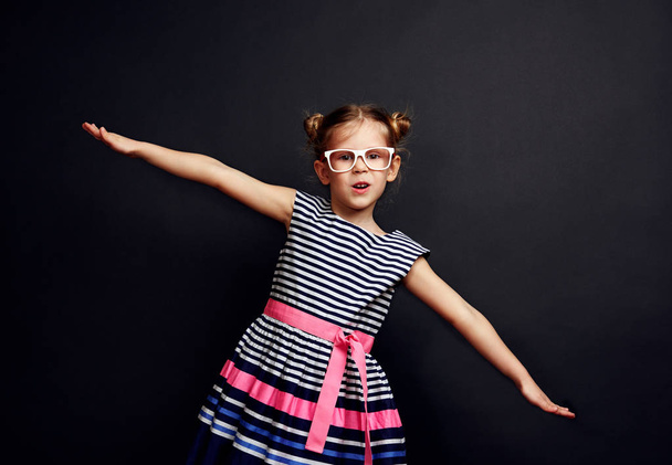 Portrait of charming girl imagining airplane fly. Cute kid in stylish dress and glasses over studio background. - Fotoğraf, Görsel