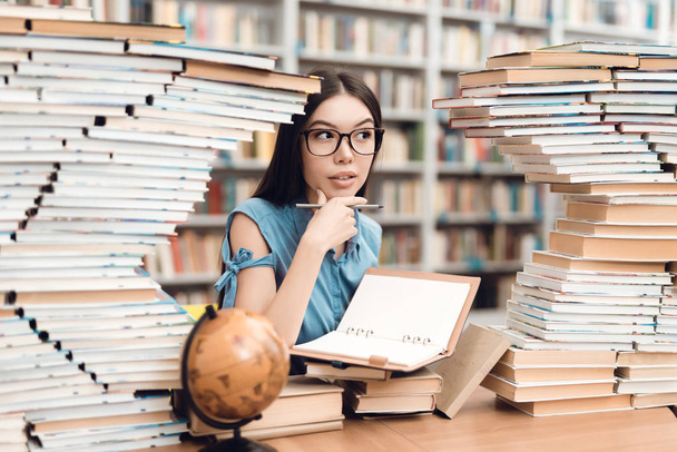 Ethnic asian girl in glasses sitting at table surrounded by books in library and writing in notebook - Photo, Image