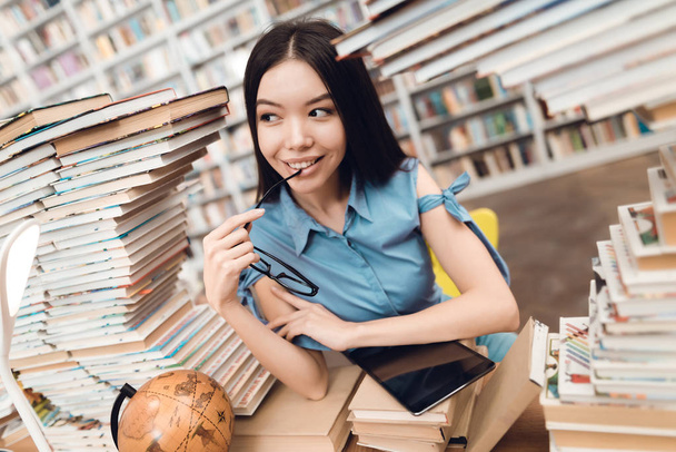 Ethnic asian girl sitting at table surrounded by books in library and using tablet - Фото, изображение