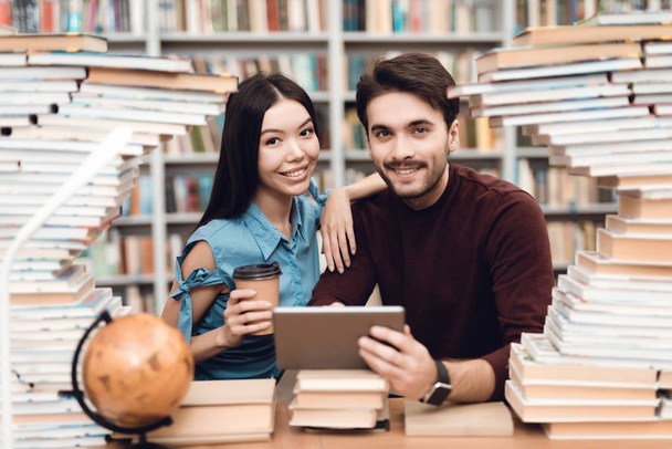 Ethnic asian girl and white guy sitting at table surrounded by books in library and using tablet - Photo, image