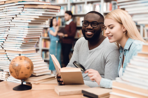 Ethnic african american guy and white girl sitting at table surrounded by books in library and reading book - Фото, изображение