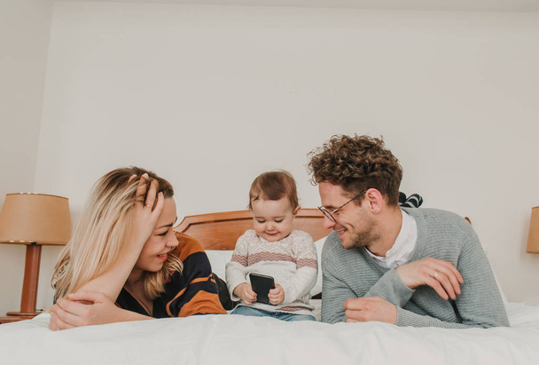 Веселая пара, лежащая в постели с маленьким ребенком, играющим со смартфоном
. - Фото, изображение