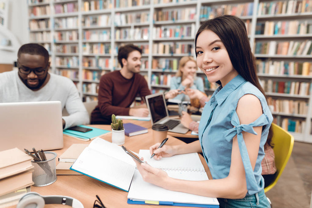 Group of ethnic multicultural students sitting at table in library, asian girl taking notes - Photo, Image