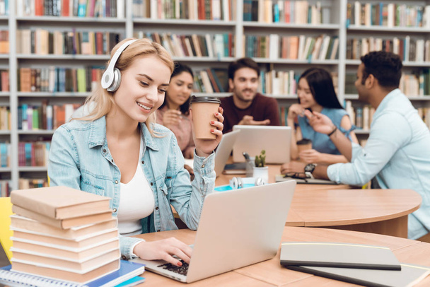 Группа этнических мультикультурных студентов, сидящих за столом в библиотеке, белая девушка, работающая на ноутбуке с наушниками
 - Фото, изображение