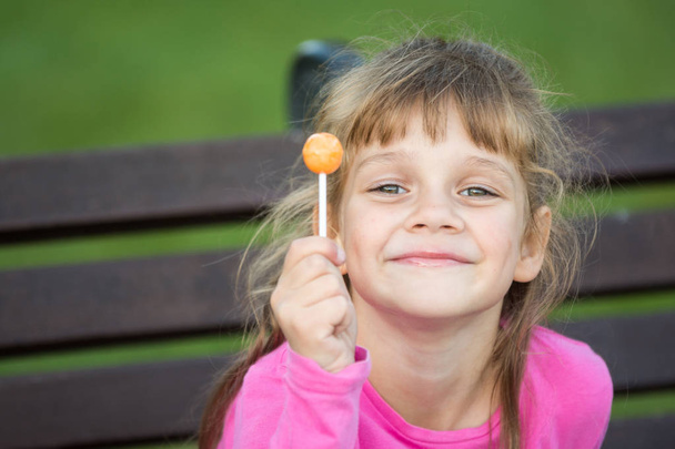 Retrato de una niña alegre de seis años que sostiene una piruleta en la mano y mira complacida al marco
 - Foto, Imagen