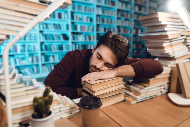 White guy sitting at table surrounded by books in library and sleeping - Foto, afbeelding