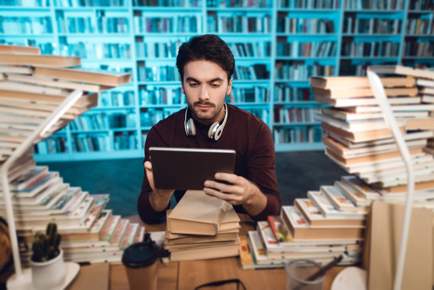 weißer Kerl sitzt am Tisch, umgeben von Büchern in der Bibliothek und mit Tablet - Foto, Bild