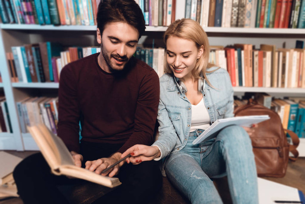 White guy and girl sitting near bookshelves in library reading books - Фото, изображение