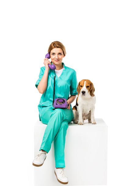 χαμογελώντας κτηνίατρος με σκύλο Μπιγκλ κοντά μιλώντας στο τηλέφωνο που απομονώνονται σε λευκό - Φωτογραφία, εικόνα