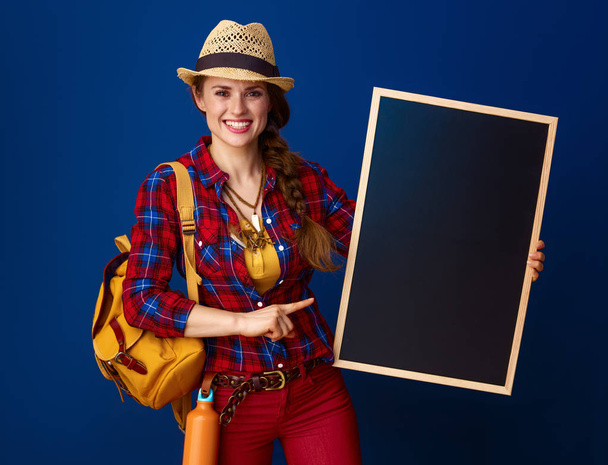smiling healthy woman hiker with backpack pointing at blank board  - Fotoğraf, Görsel