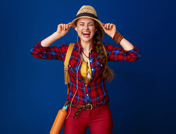 Searching for inspiring places. smiling fit woman hiker in a plaid shirt with headphones listening to the music pulling a hat on blue background - Fotografie, Obrázek