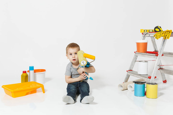 Little cute chłopiec siedzi na podłodze z instrumentów do remontu mieszkanie Pokój na białym tle. Tapety, klejenia akcesoria, narzędzi do malowania. Naprawa główna. Rodzicielstwa, koncepcja dzieciństwa. - Zdjęcie, obraz