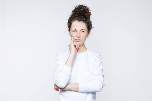 Young American female in white t-shirt looking at the camera with serious pensive expression, touching her cheek with index finger, posing against studio wall with copy space for your information. - Photo, Image