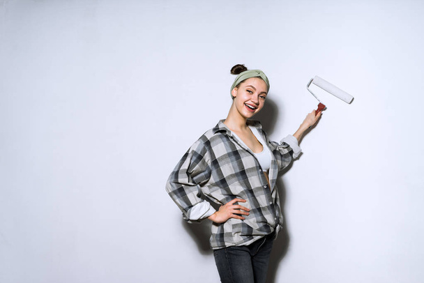 happy young girl in a plaid shirt doing repairs in her new apartment, painting the walls with a roller - Foto, Bild