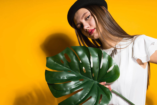 confident stylish girl model in a black hat holds a green leaf, posing on a yellow background - Foto, afbeelding