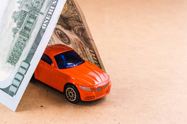 Close-up hundred dollar bill roof covers red car toy on brown background. Concept of car insurance - Фото, изображение