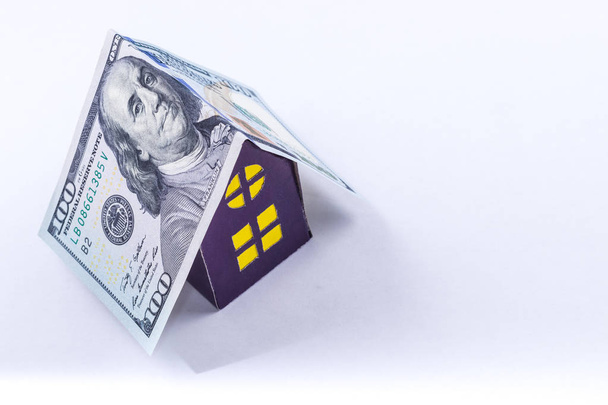 hundred dollar bill roof covers purple cardboard house with yellow windows isolated on white background.Concept of a good investment - Photo, Image