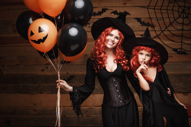 Halloween Witch Concept - madre alegre y su hija en trajes de bruja celebrando Halloween posando con globo naranja y negro sobre murciélagos y tela de araña sobre fondo de estudio de madera
. - Foto, Imagen