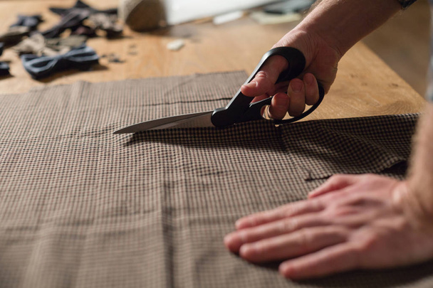 Closeup snijden bruine geruite wollen stof. het lijnpatroon. Jonge man werken als een kleermaker en een naaimachine gebruiken in werkplaats. - Foto, afbeelding