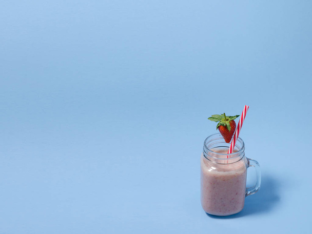 Roze aardbei smoothie in een glas mason jar met stro en verspreide bessen op blauwe achtergrond. Gezond en lekker ontbijt. Biologische aardbei Smoothie gemaakt met verse ingrediënten. - Foto, afbeelding