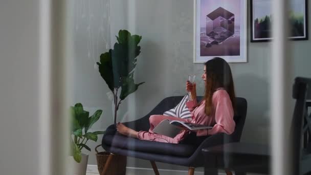 Uma linda garota de pijama rosa sexy lê uma revista brilhante deitada no sofá e bebendo champanhe de um copo
. - Filmagem, Vídeo