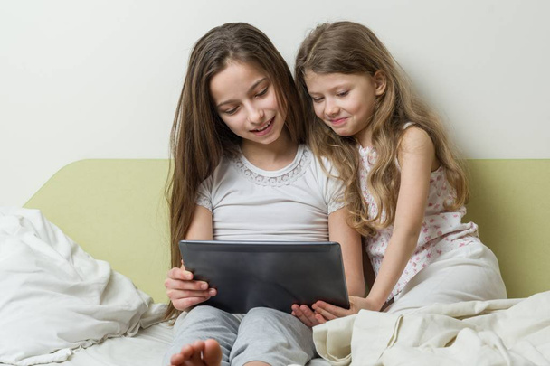 Two girls in pajamas are sitting in bed at home with interest looking at the tablet and laughing, chatting - Фото, изображение