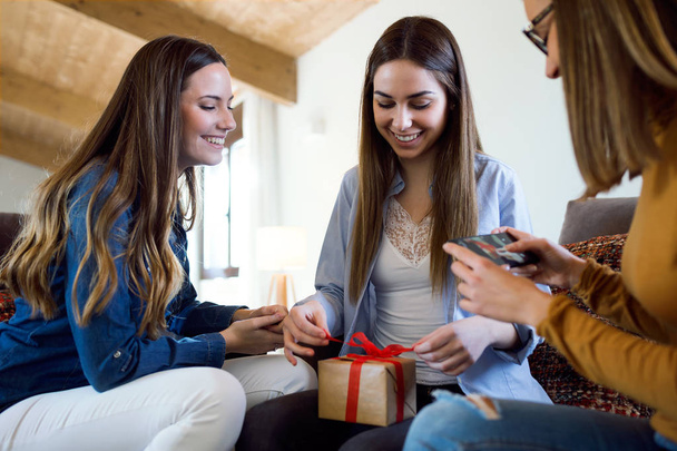 Two beautiful young women exchanging a gift while their friend takes a photo at home. - Foto, afbeelding