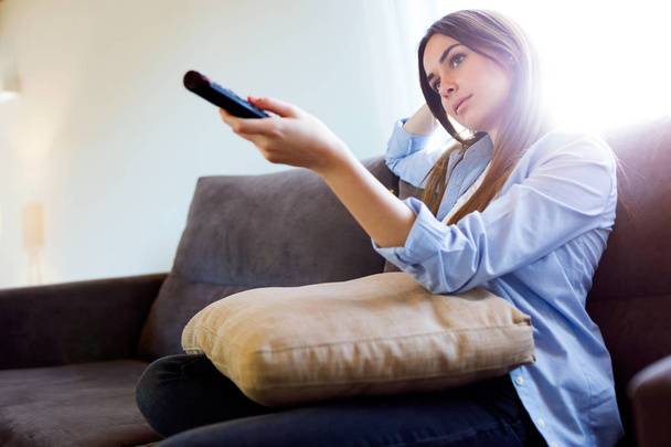 Beautiful bored young woman watching TV and holding remote control at home. - Photo, Image