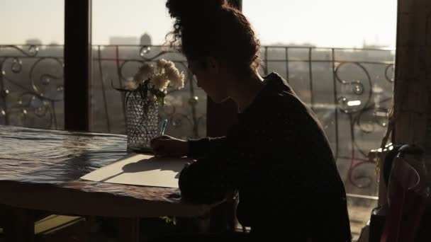 Vrouwelijke artiest zit door de tabel met bloemen erop, op papier tekenen met pen, in een zeer minimalistische kunststudio op de achtergrond. - Video