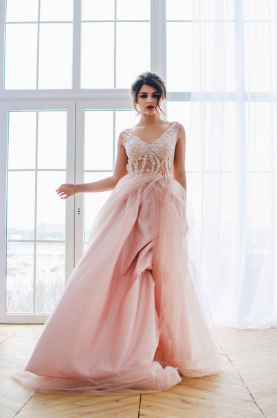 Mooi meisje in een luxe roze jurk is permanent in de buurt van het venster - Foto, afbeelding