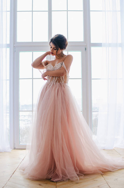 Menina bonita em um vestido rosa luxuoso está de pé perto da janela
 - Foto, Imagem