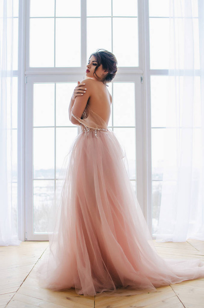 Σε ένα πολυτελές ροζ φόρεμα το όμορφο κορίτσι στέκεται κοντά στο παράθυρο - Φωτογραφία, εικόνα