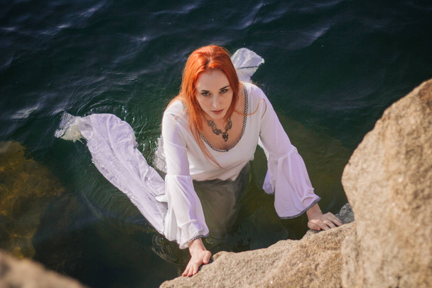 Menina ruiva bonita está flutuando na água. Costa rochosa. Um modelo em água esmeralda em um vestido antigo branco. Imagem medieval. Fantasia
 - Foto, Imagem