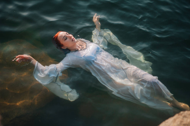 Beautiful red-haired girl is floating in the water. Rocky coast. A model in emerald water in a white antique dress. Medieval image. Fantasy - Photo, Image