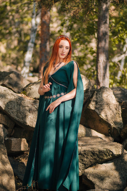 ターコイズ ブルーのドレスと彼女の頭の上の王冠の美しい赤い髪の少女は、秋の森で歩いています。エルフのイメージ。山の中の大きな岩です。太陽のまぶしさ。ファンタジー - 写真・画像