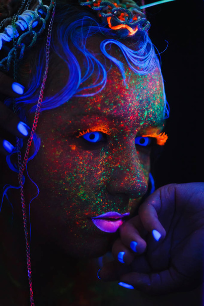 Νέον πορτρέτο ενός κοριτσιού. Bright πιτσιλιές στο πρόσωπο. Μοντέλο με μπλε νέον φακούς στο σκοτάδι. Η εικόνα για τις Απόκριες. - Φωτογραφία, εικόνα