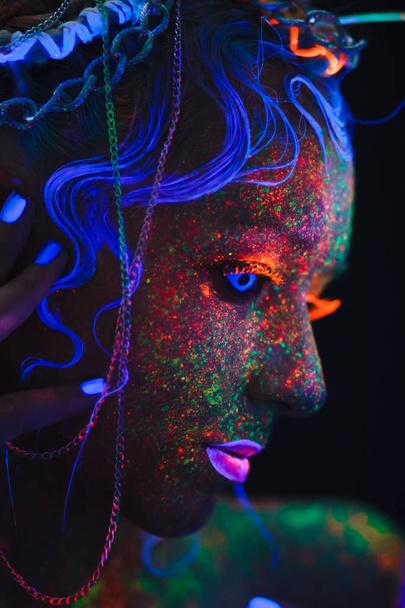 Νέον πορτρέτο ενός κοριτσιού. Bright πιτσιλιές στο πρόσωπο. Μοντέλο με μπλε νέον φακούς στο σκοτάδι. Η εικόνα για τις Απόκριες. - Φωτογραφία, εικόνα