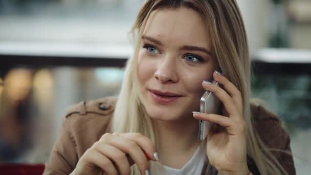 Pretty blonde woman talks on the phone sitting in the restaurant and drinking a milkshake - Záběry, video