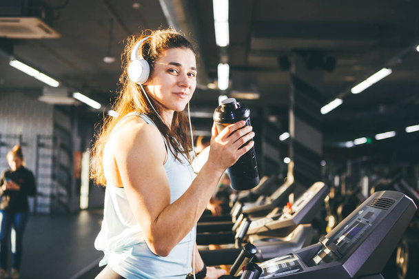 Sports nutrition and exercise. beautiful woman holds protein in her hand in bottles, shaker, listens music in headphones on head in gym running on treadmill, at hands of sports watch,in sunny weather - Photo, Image