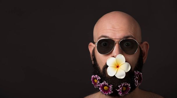 neoholený muž s vousy s květinami a sluneční brýle na tmavém pozadí s copyspace - Fotografie, Obrázek