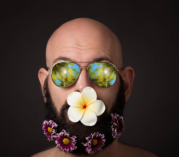 borostás férfi szakállal, virágokkal és napszemüveg sötét háttér - Fotó, kép