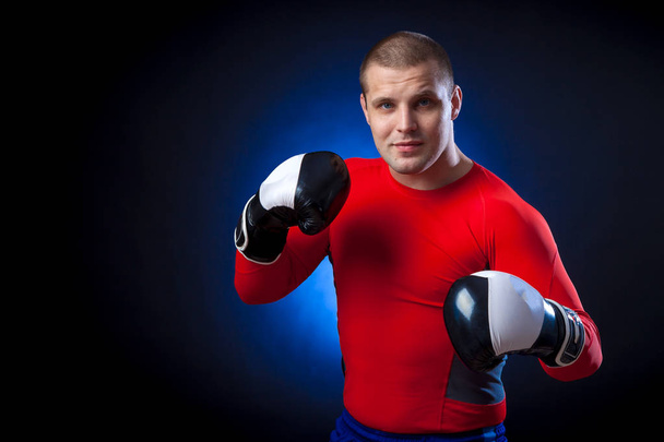 Un boxeur sportif aux cheveux foncés fort dans un garde-robe de sport rouge et dans des gants de boxe noir et blanc boxant contre un bleu une lumière sur un fond isolé noir
  - Photo, image