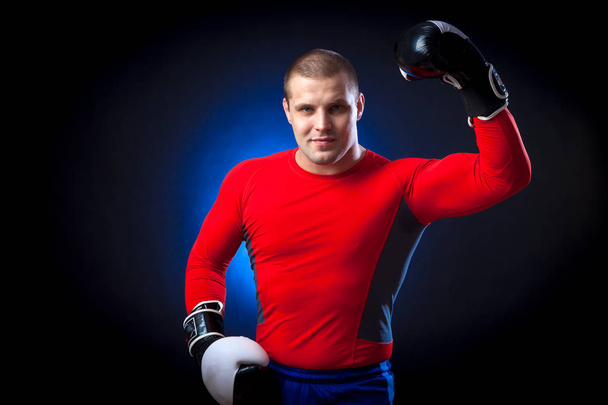 ein kräftiger dunkelhaariger Boxer in roter Sportkleidung und schwarz-weißen Boxhandschuhen lächelt und steht vor blauem Licht auf schwarzem Hintergrund  - Foto, Bild