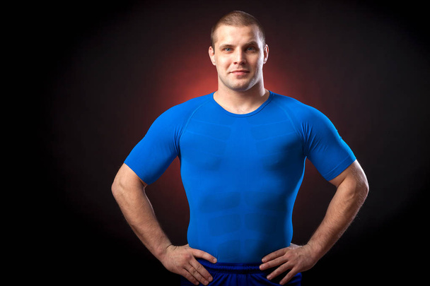 A strong dark-haired sportman  in a blue sports wear  rush guard posing against a red a lights on a black isolated  background  - Photo, Image
