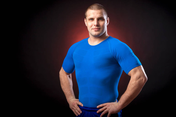 A strong dark-haired sportman  in a blue sports wear  rush guard posing against a red a lights on a black isolated  background  - Photo, Image