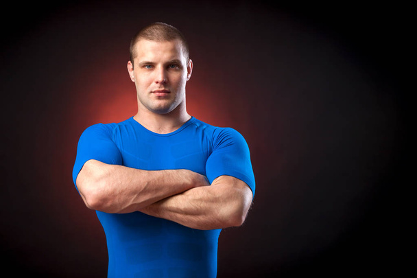 A strong dark-haired sportman  in a blue sports wear  rush guard  looks seriously at the camera and holds his arms crossed on his chest against a red a lights on a black isolated  background  - Photo, Image